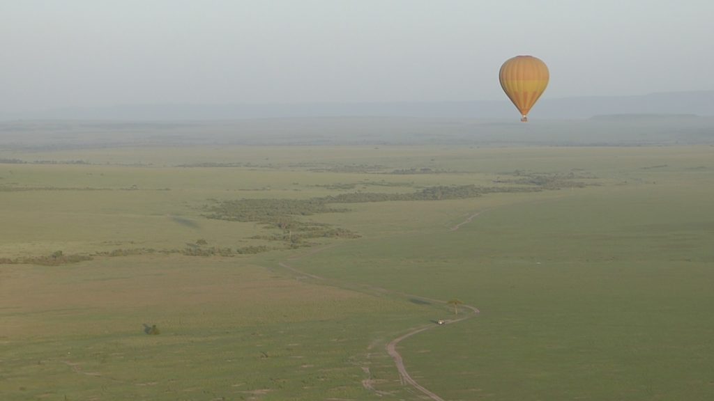 The Maasai Mara vom above. A small street is visible on the ground, in the distance you see an area full of trees. A yellow and orange balloon is flying in front.