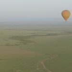 The Maasai Mara vom above. A small street is visible on the ground, in the distance you see an area full of trees. A yellow and orange balloon is flying in front.