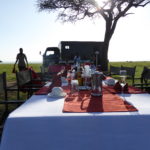 A breakfast table filled with champagne and coffee. In the back you see a truck and the small kitchen of the Balloon Safari where a chef is preparing food.