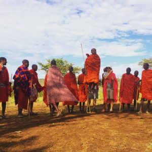 Maasai men in red shukas are performing their jumping dance. One man is jumping very high infront of the others watching. Two visitors are amazed by the performance.