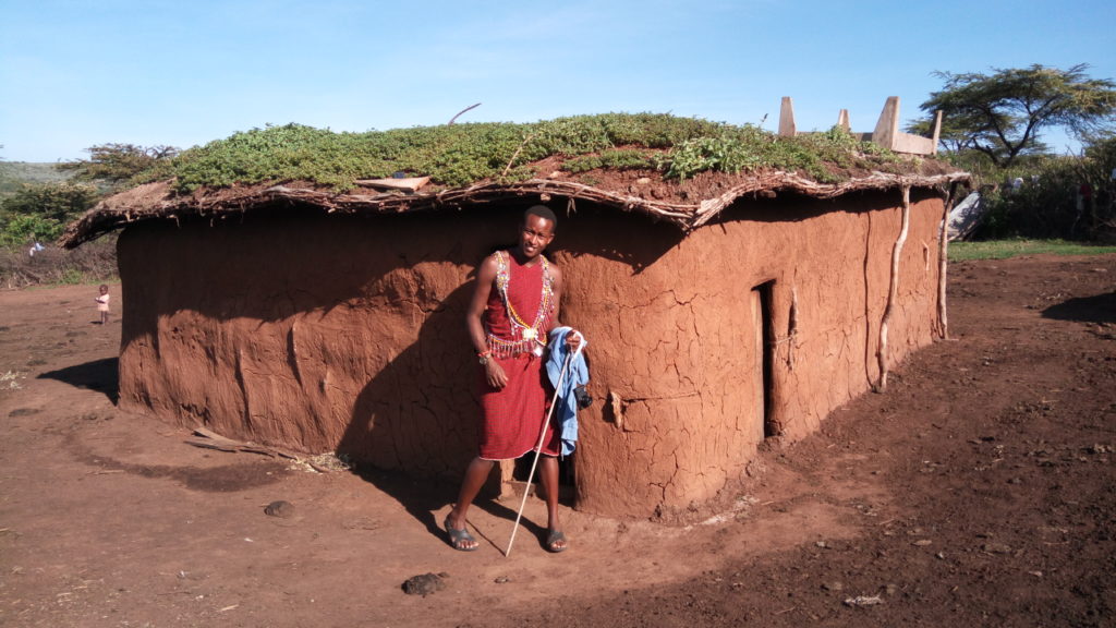 Experience Maasai culture in Kenya. Maasai man standing in front of a typical Maasai house out of mud, cow dung and clay. Gras is grwoing on the roof.