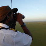 A pilot in a white shirt is looking though his binoculars. He is on the lookout to find animals down on the ground. You see another balloon in the far distance