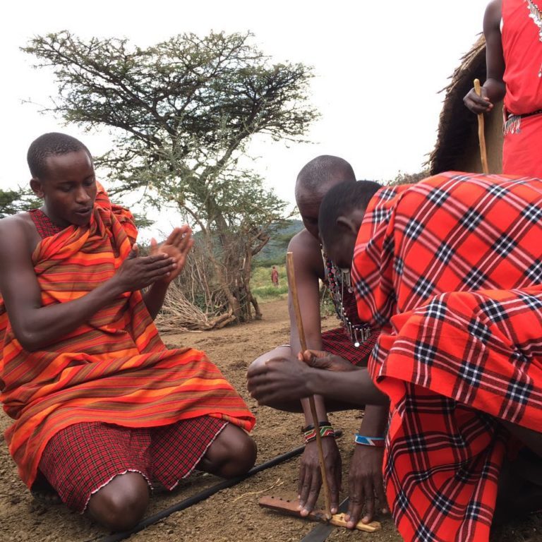 Three Maasai men making fire with the traditional two woods.