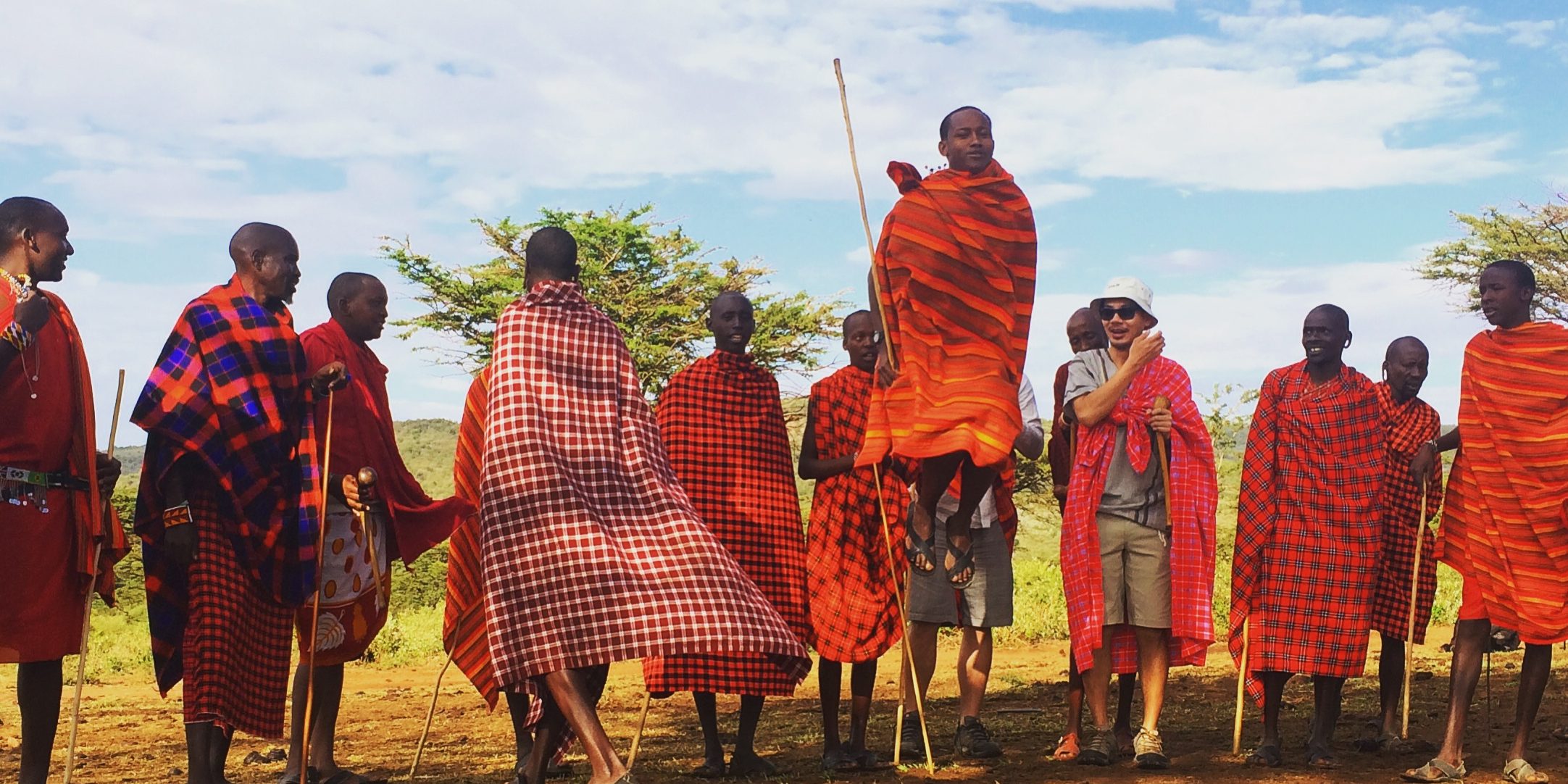 Maasai men in red shukas are performing their jumping dance. One man is jumping very high infront of the others watching. Two visitors are amazed by the performance.